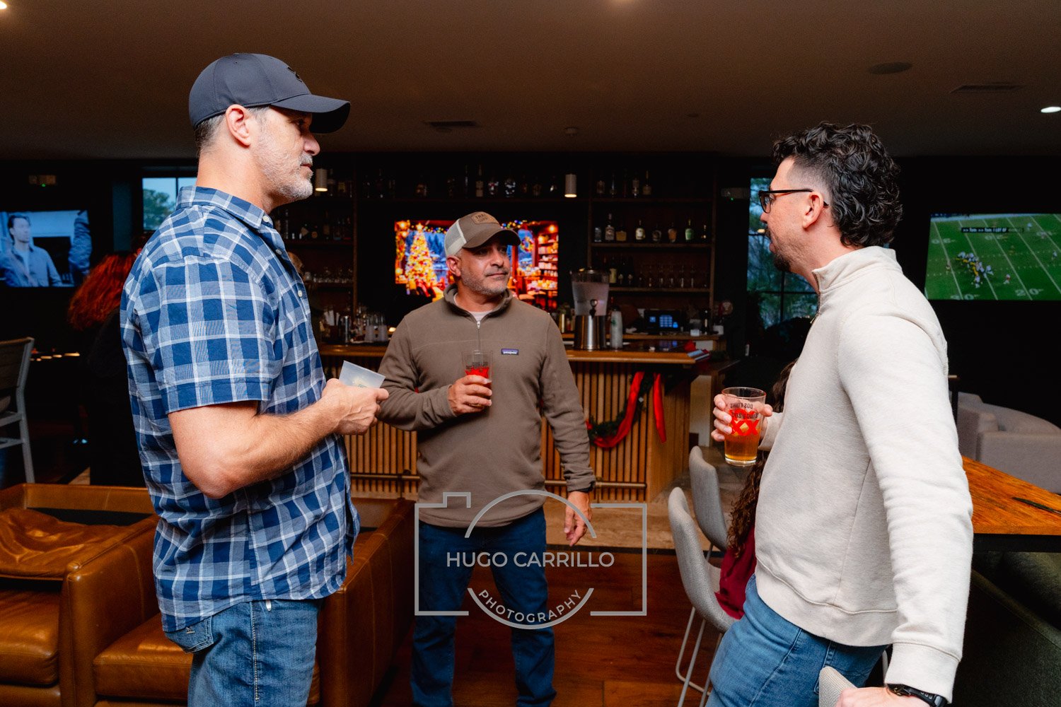 Three men are conversing at a bar, each holding a drink. The man on the left wears a blue checkered shirt and a dark hat, the man in the middle wears a brown zip-up jacket and a baseball cap, and the man on the right wears a white quarter-zip sweater
