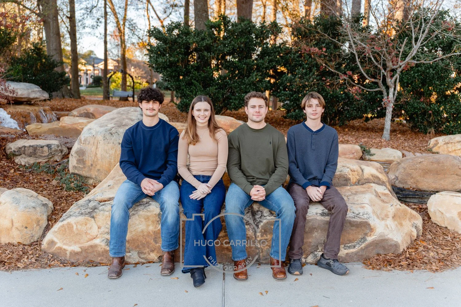 Four young people sitting on a large rock in a park, with trees and foliage in the background, during autumn.
