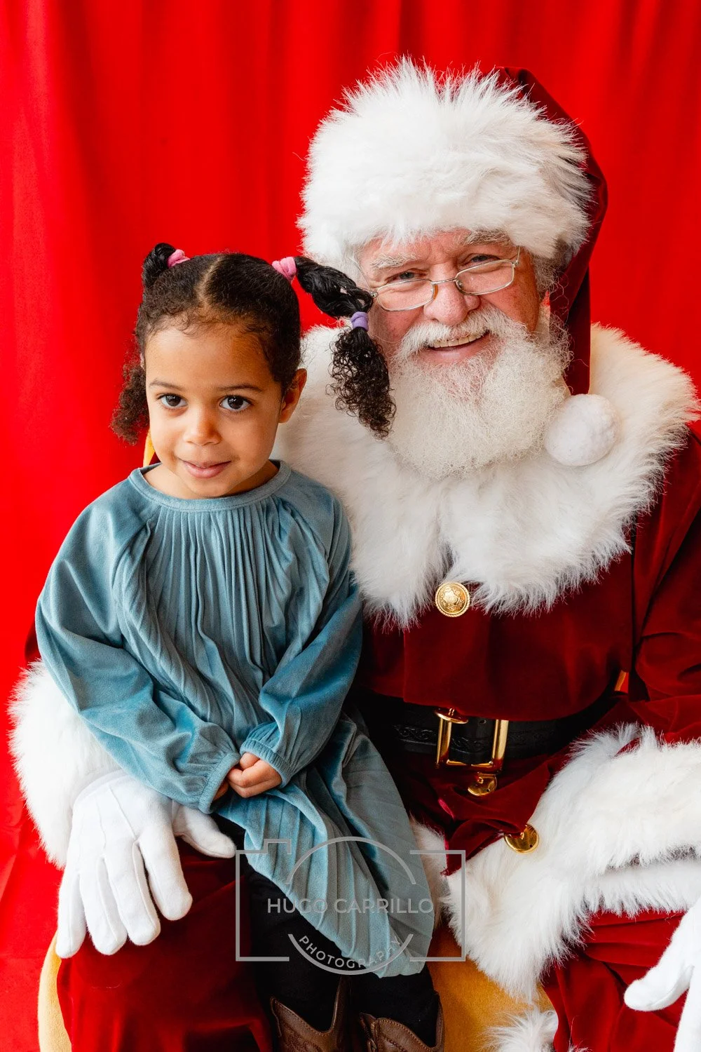 Young girl with curly hair in pigtails sitting on Santa Claus's lap next to him with a red curtain background.