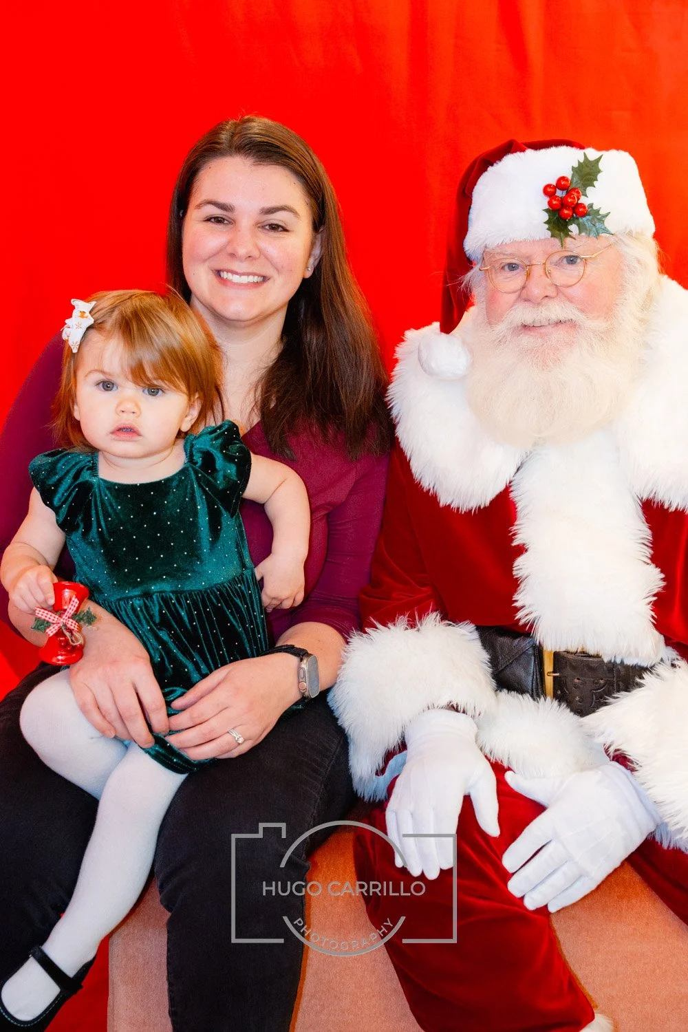 A woman and a young girl sitting next to Santa Claus dressed in a red suit with white fur trim. The woman and girl are smiling, and the girl has a white bow in her hair and is holding a small red holiday ornament. The background is red.