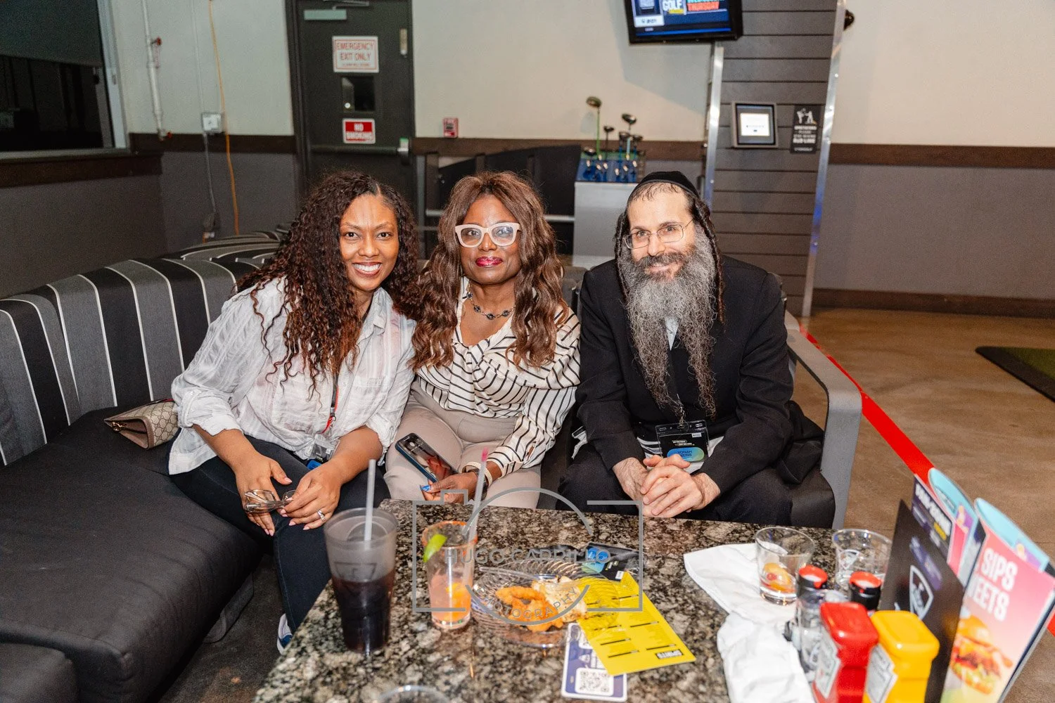 Three people sitting together at a table in a restaurant or bar, smiling at the camera. The table has drinks, menus, and condiments.