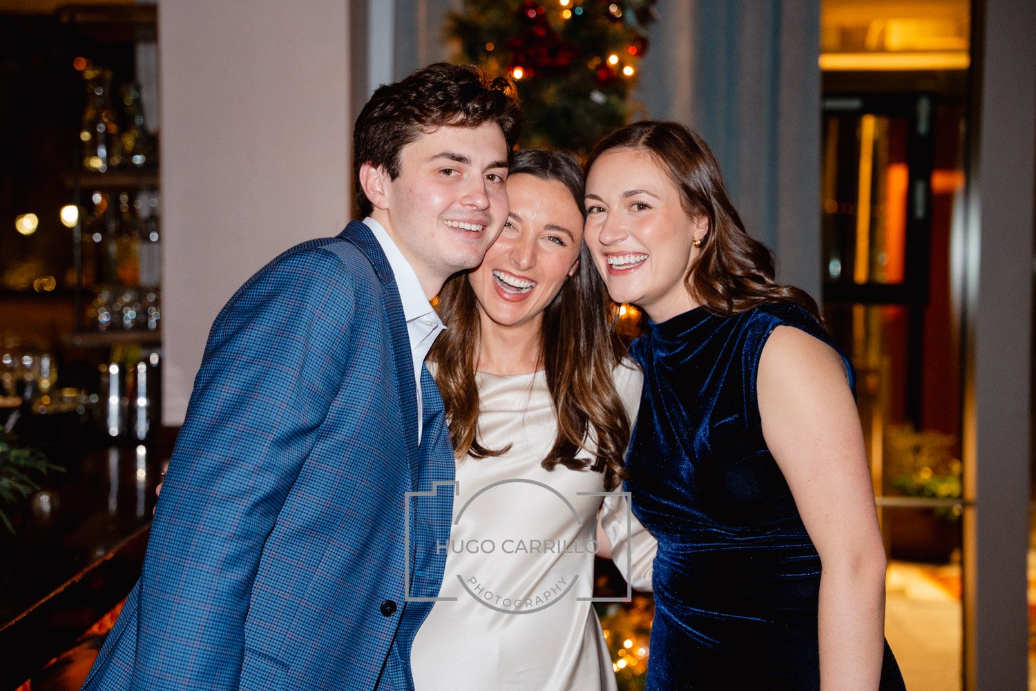 Three people smiling and posing together indoors during a holiday celebration, with a decorated Christmas tree in the background.