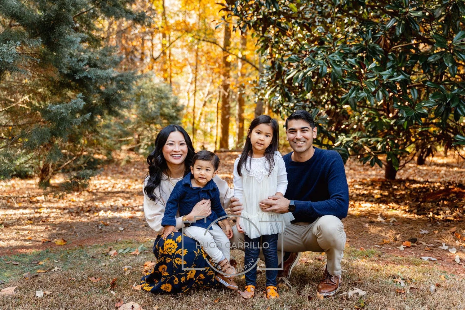 A family of four in a wooded autumn park with fall foliage, smiling at the camera. The mother has long black hair, the father has short black hair, the daughter has long dark hair, and the young son has short black hair. The mother is wearing a light