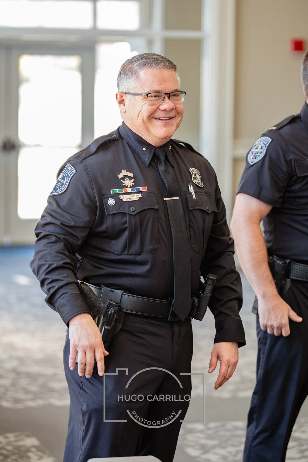 A smiling police officer in uniform standing indoors near a window.