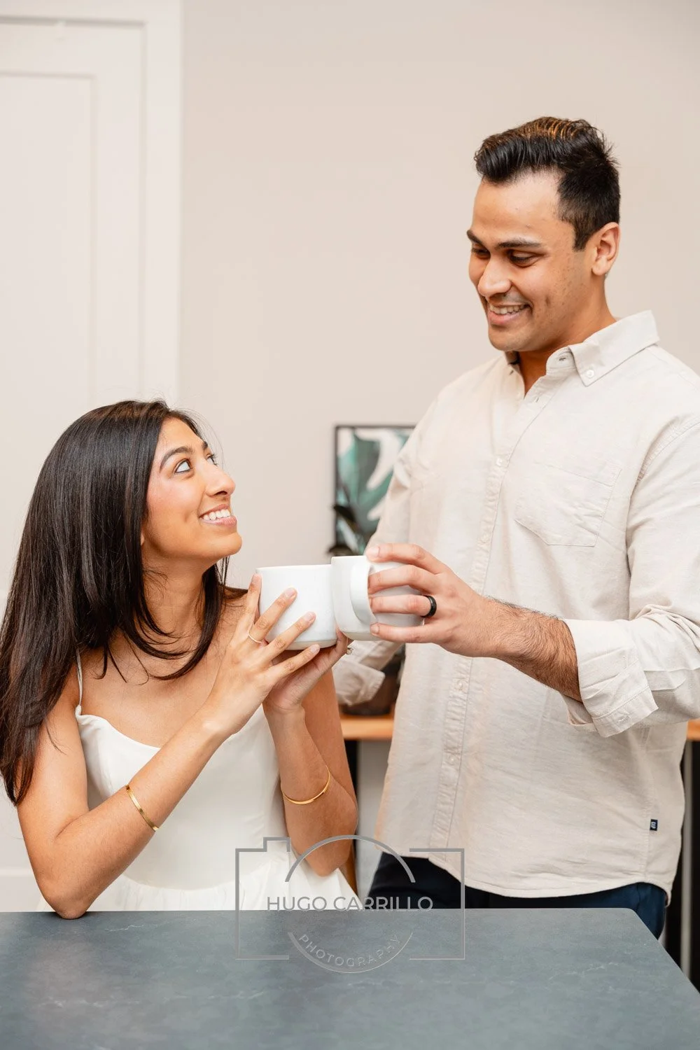 A woman sitting at a table holding a white coffee mug, smiling up at a man who is handing her another mug, both are smiling, in a bright indoor setting.