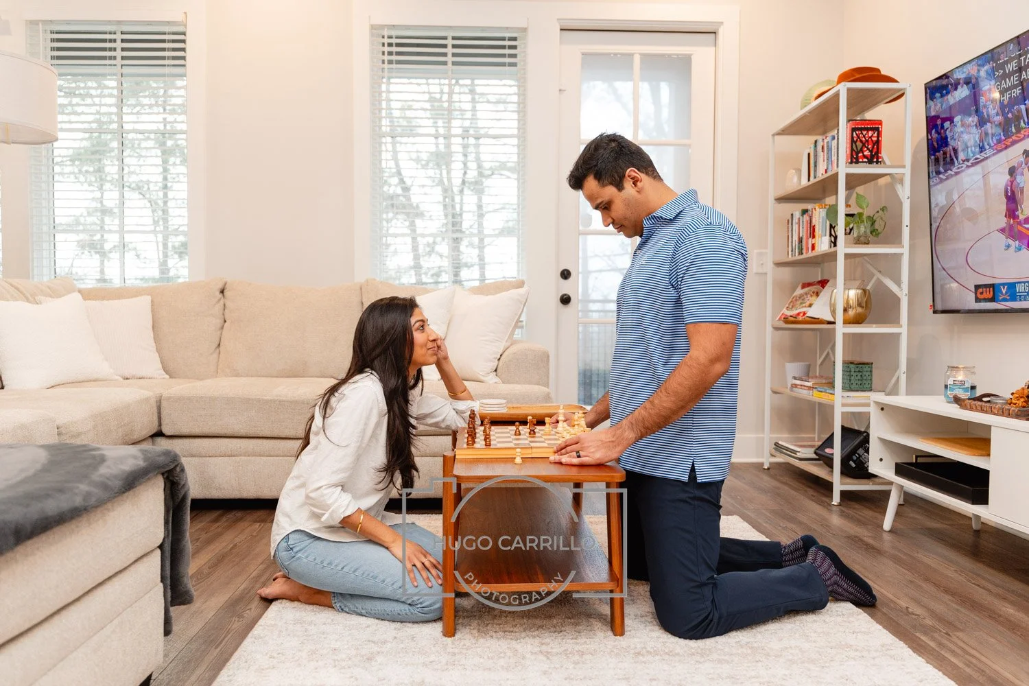 A man and woman playing chess on a coffee table indoors, the woman sitting on the floor, the man kneeling, in a living room with beige couches, white shelves, and a large flat-screen TV.