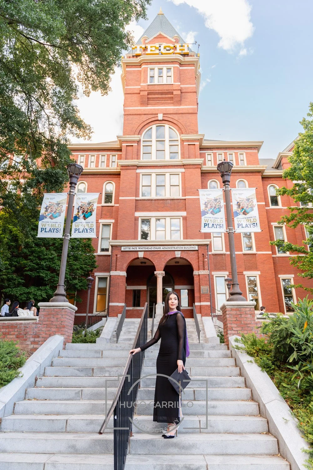 A woman in a black dress standing on the steps leading to Lettie Pate Whitehead Evans Administration Building, a red brick historic university building with a clock tower, surrounded by trees and banners that say "Welcome to where the world plays!".