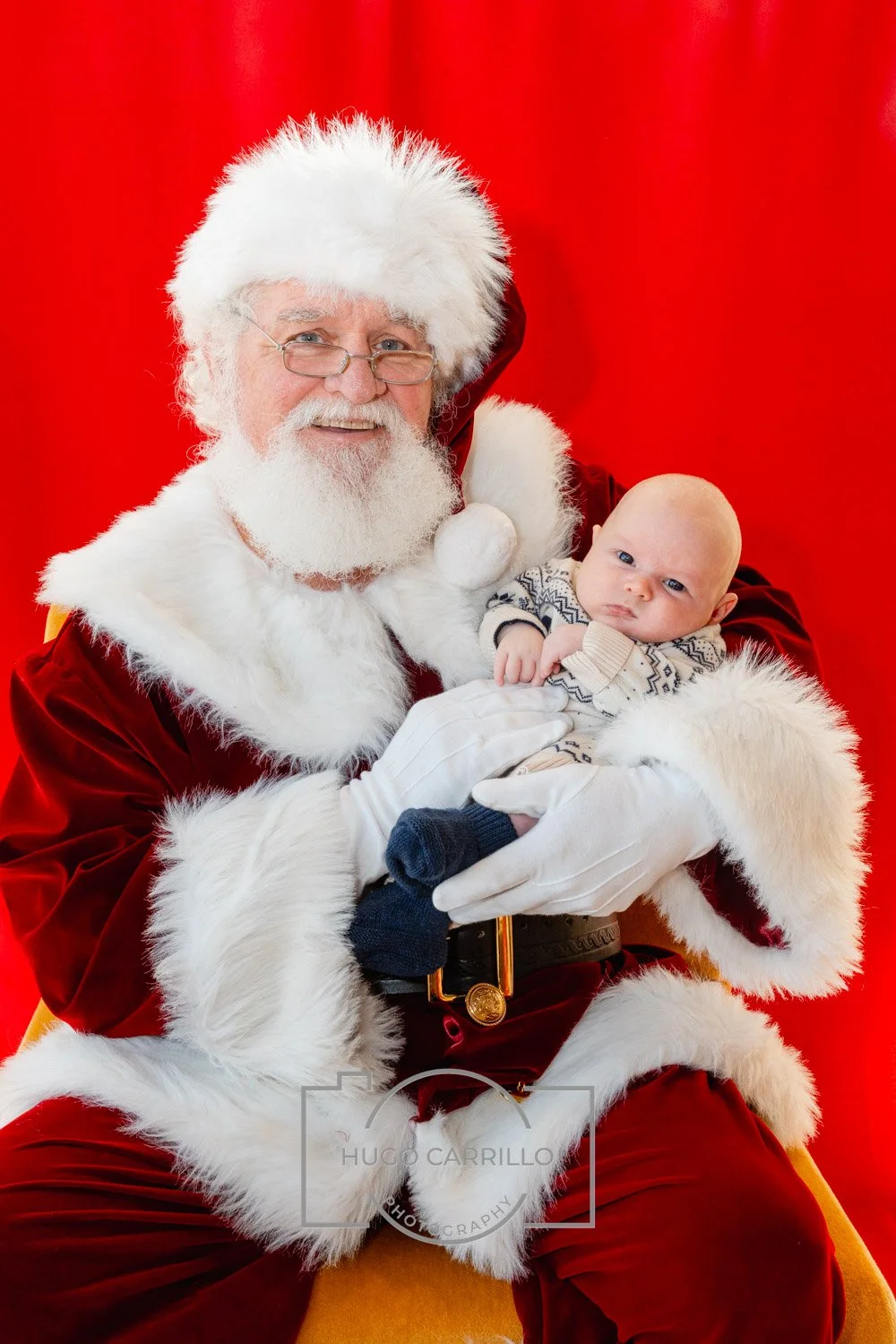 Santa Claus holding a baby in front of a red background.