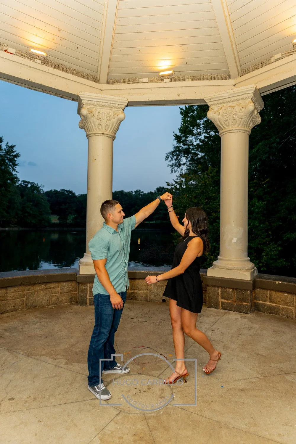 A young man and woman are dancing together under a pavilion by a river at dusk. The man is wearing a turquoise shirt and jeans, while the woman is wearing a black dress and high heels. They are holding hands with their arms raised, and the pavilion h