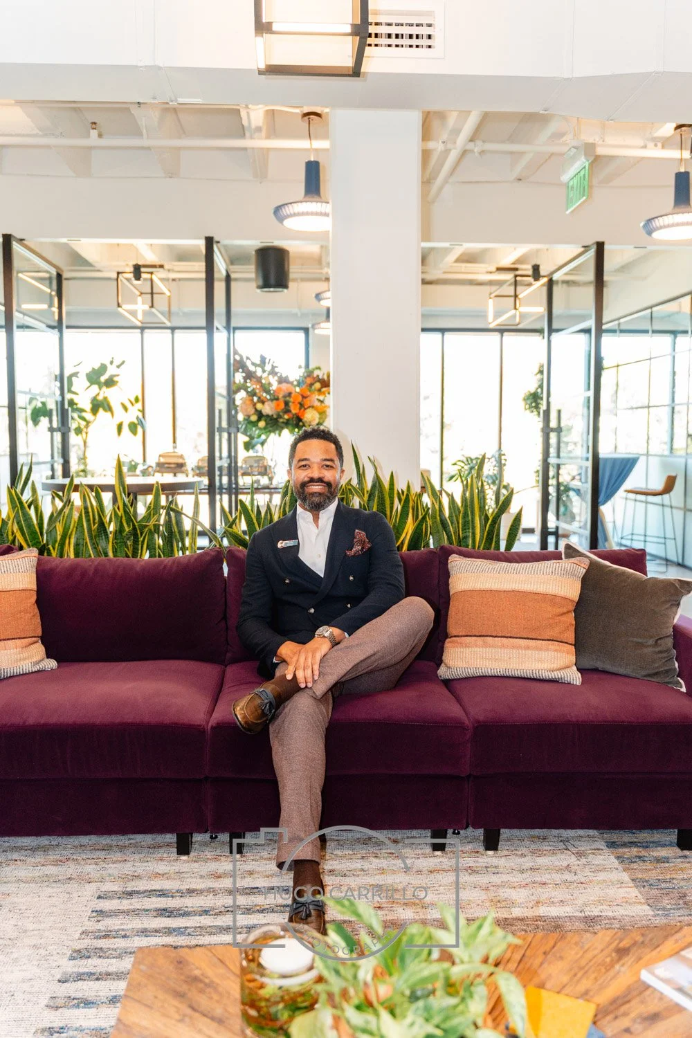 A man with a beard and mustache, wearing a dark blazer, white shirt, and gray pants, sitting on a maroon sofa in a bright, modern indoor space with plants and large windows.