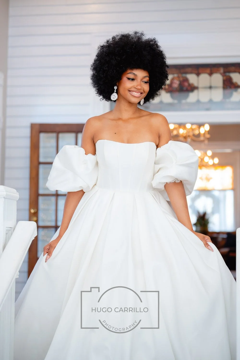 A woman in a white wedding dress with puffed sleeves, holding the sides of her dress, smiling indoors with chandeliers and a wooden door in the background.
