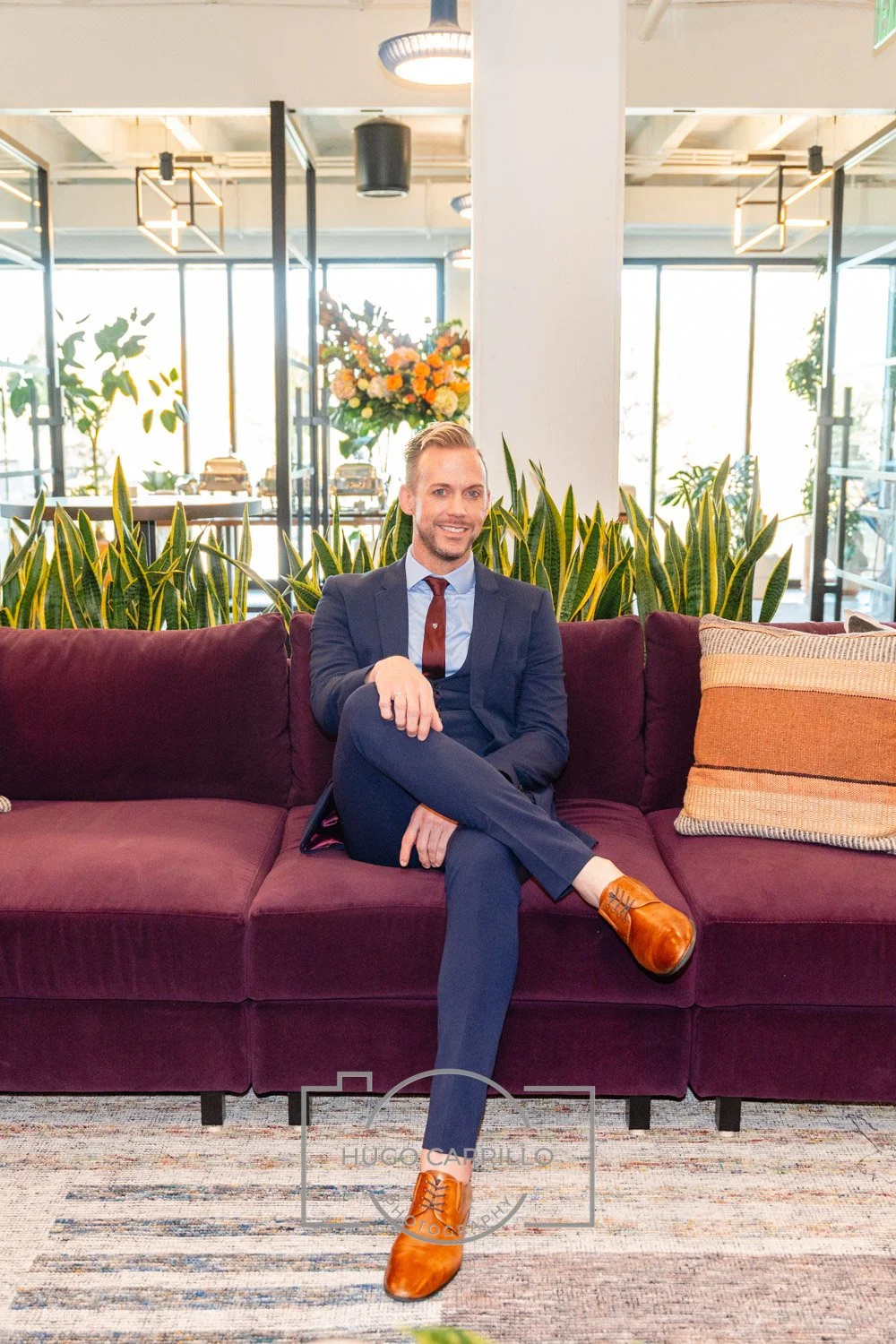 A man sitting on a burgundy sofa in a modern, well-lit space with large windows, green plants, a bouquet of orange and yellow flowers, and decorative pillows.