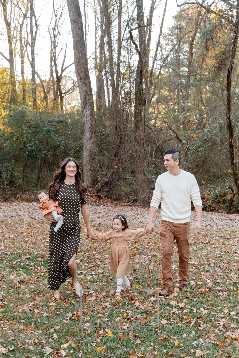 Family of four enjoying a walk in a park during autumn, holding hands, surrounded by fallen leaves and trees.