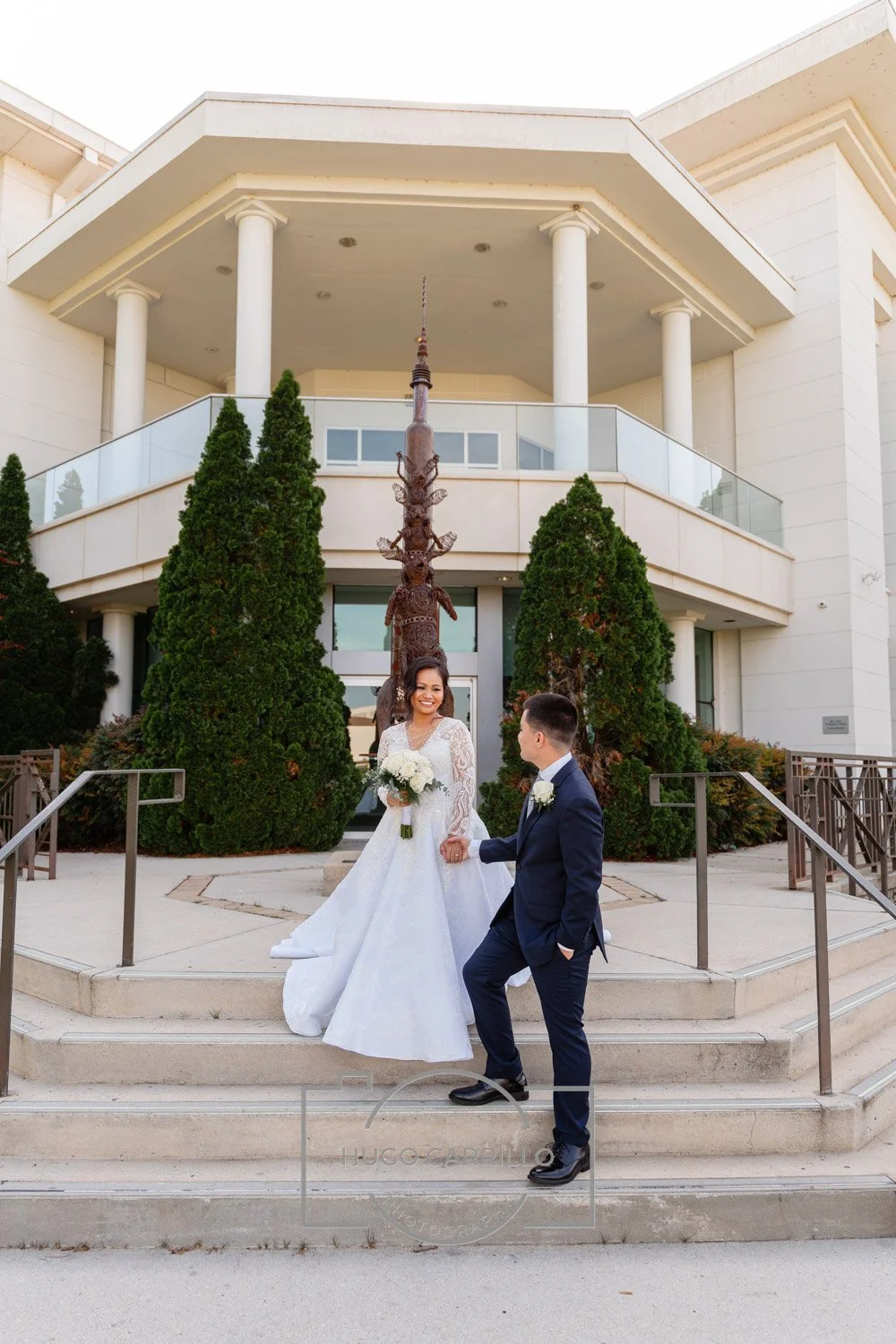 A bride and groom are standing on steps outside a modern white building with a sculpture and tall trees in the background. The bride is holding a bouquet of white flowers and wearing a white wedding dress. The groom is in a navy suit with a white bou
