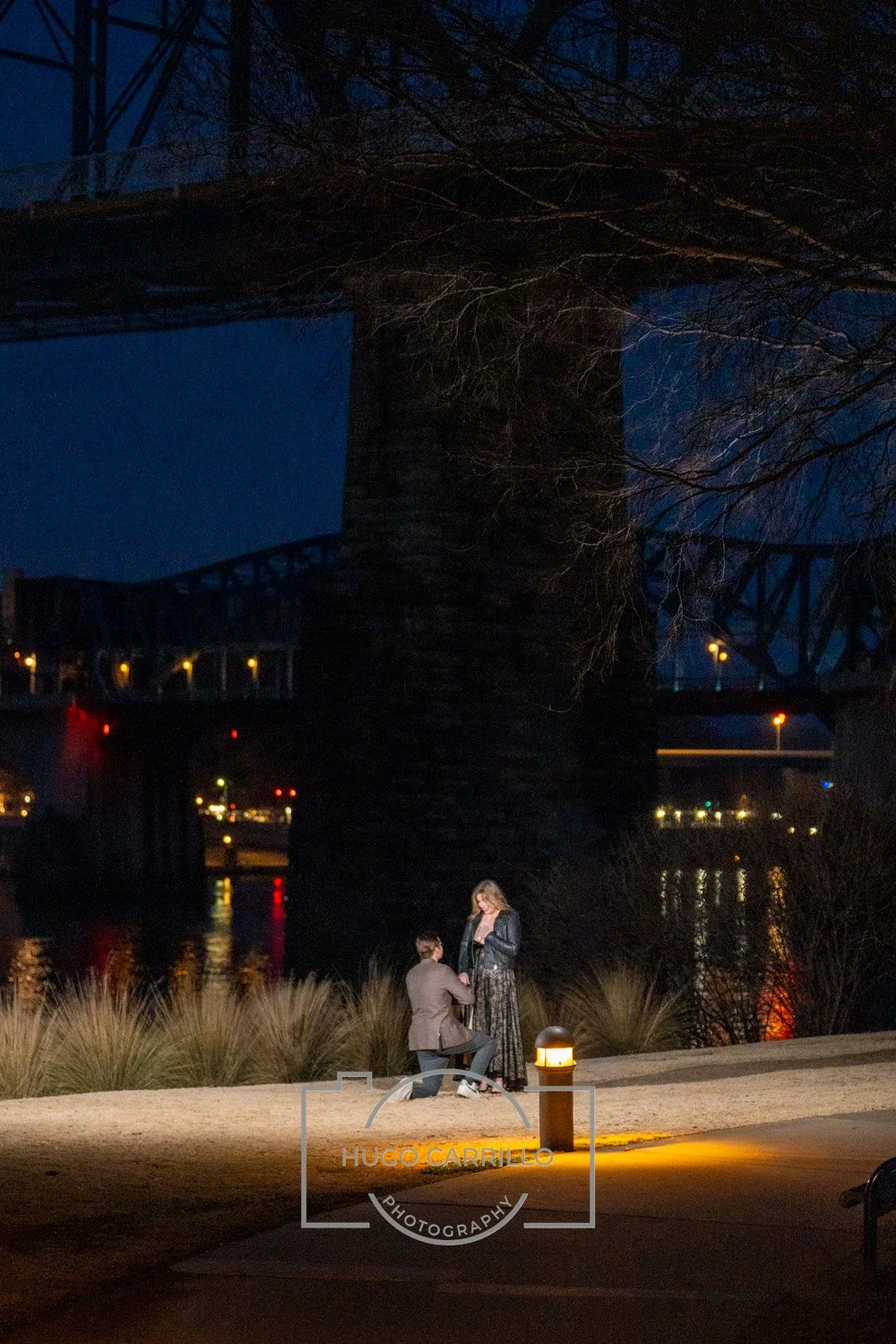 A nighttime scene of a man proposing to a woman outdoors near a river, under a large bridge with city lights reflecting on the water.