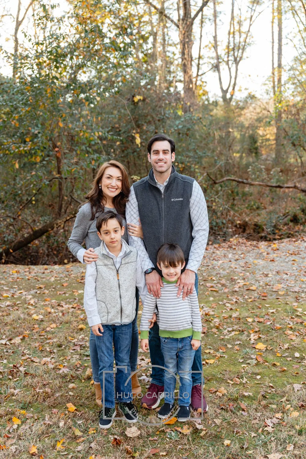 A happy family of four standing outdoors among fallen autumn leaves in a wooded area, smiling at the camera.