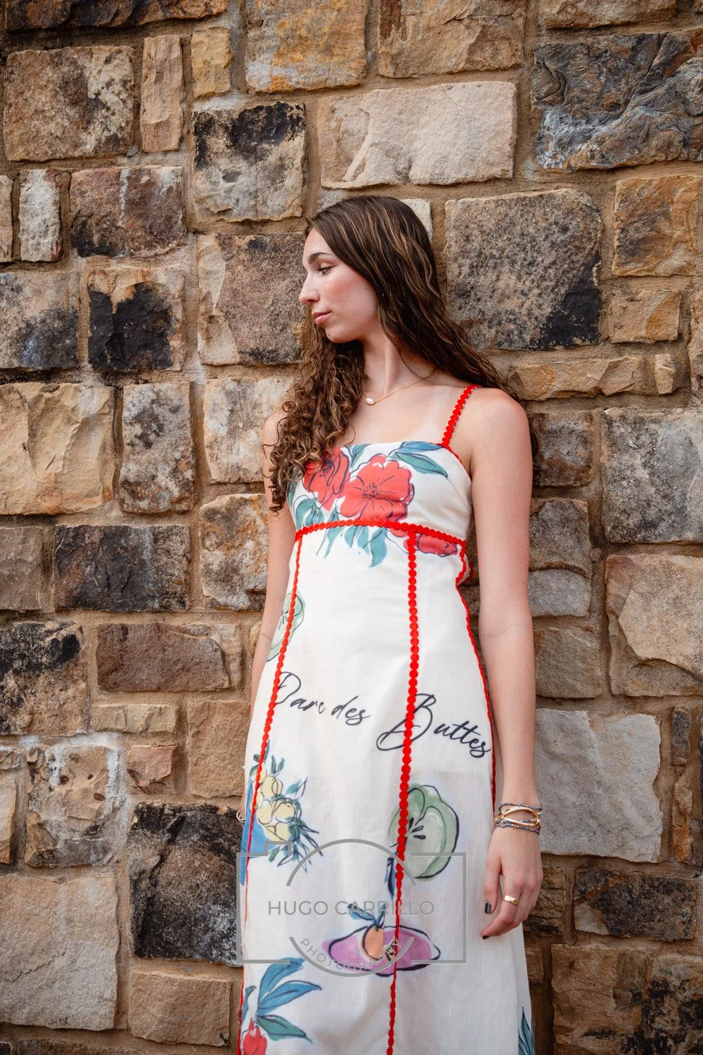A young woman with wavy brown hair wearing a white dress with colorful floral prints and red piping, standing against a stone wall.
