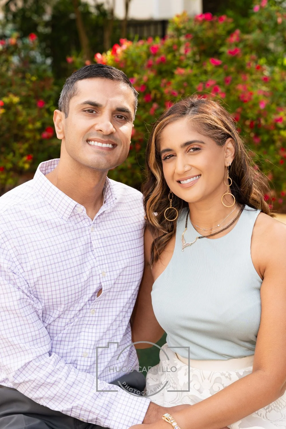 A smiling man and woman sit outdoors with pink flowering bushes and green trees in the background.