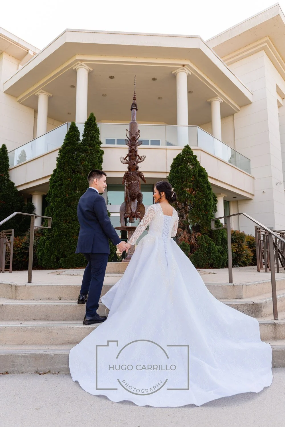 A bride and groom holding hands in front of a modern white building with a metal sculpture of a stag between green bushes.