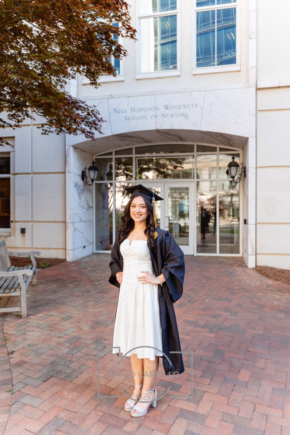 A young woman in a white dress and graduation cap and gown standing in front of the Nell Hodgson Woodruff School of Nursing building, smiling for the camera.