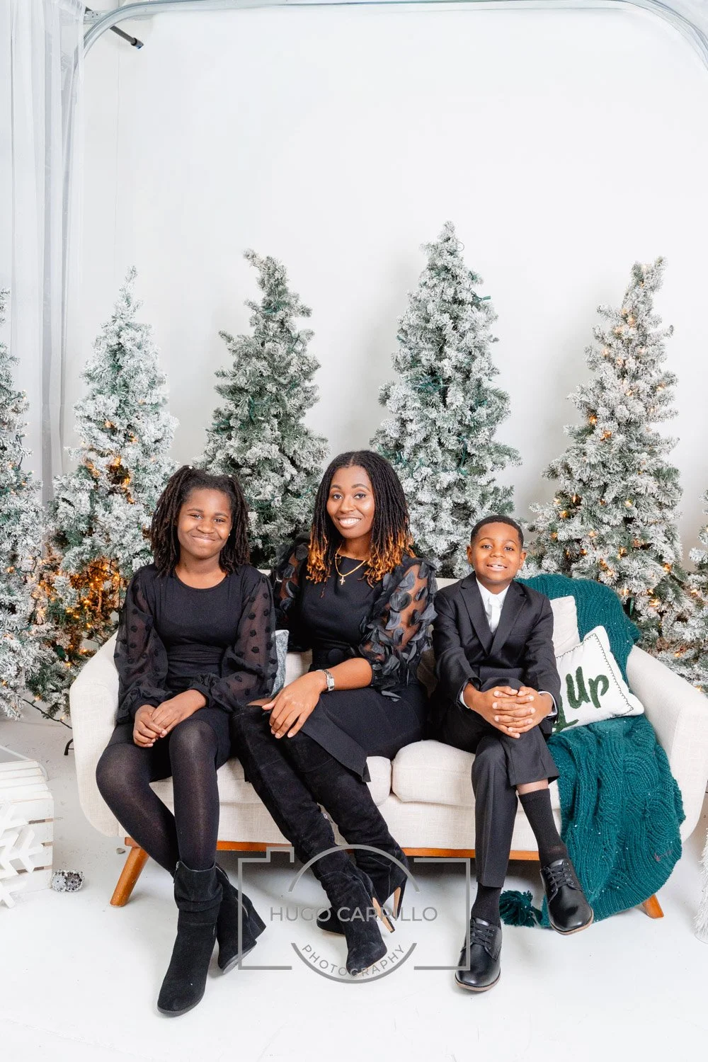 A woman and two children sitting on a white sofa with a holiday backdrop of snow-covered trees decorated with lights. The woman is in the center, wearing a black dress with sheer polka-dot sleeves, and the children on each side are in black formal at