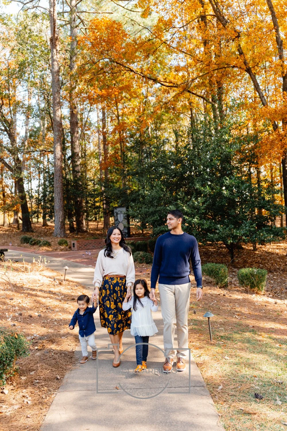 A family of four walking on a sidewalk in a park during fall. The mother and father are holding their children's hands. The mother is wearing a cream sweater and a floral skirt, and the father is dressed in a navy sweater and beige pants. The childre