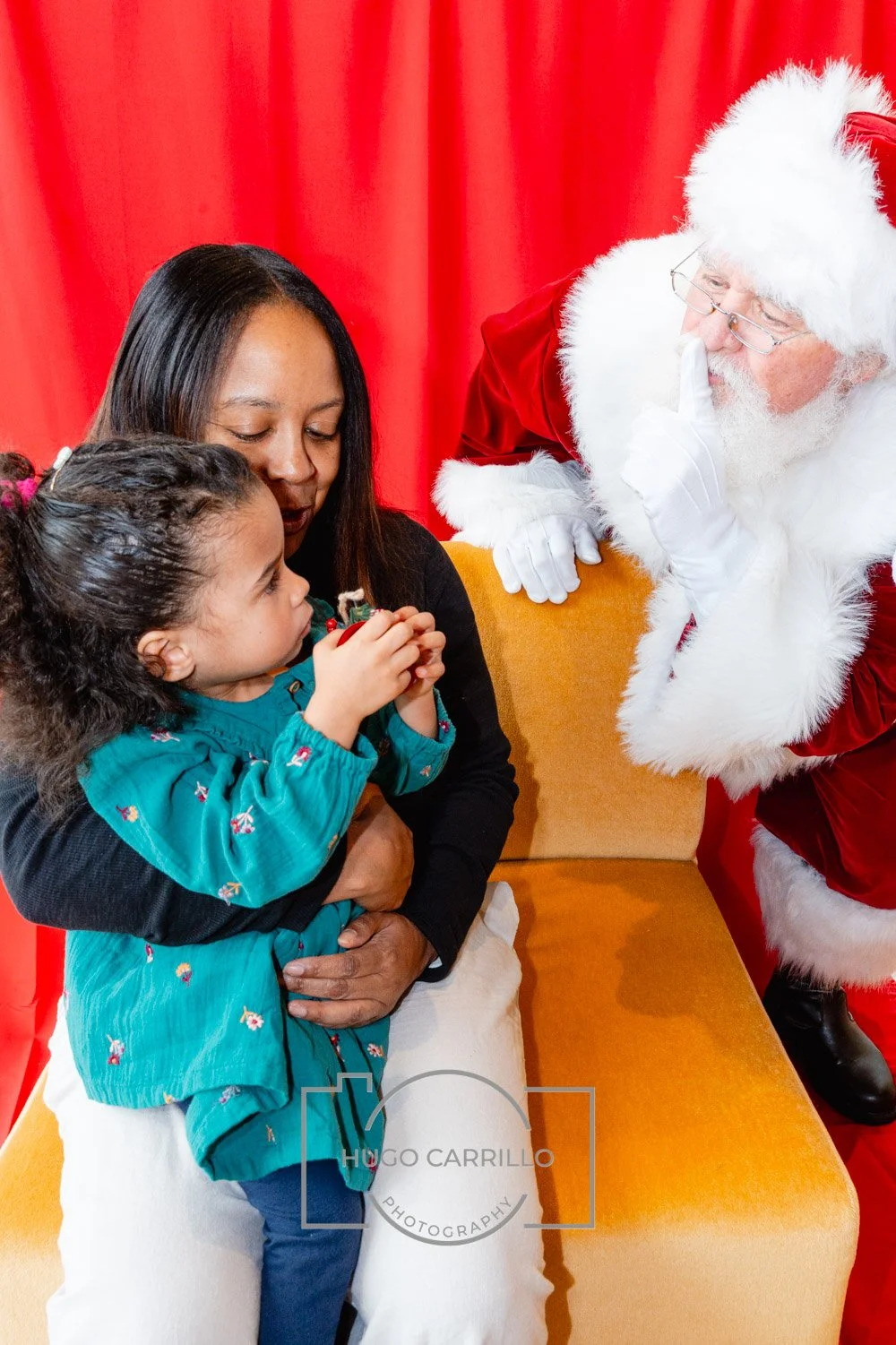 A young girl, her mother, and Santa Claus having a conversation during a Christmas event. Santa is whispering to the girl while the mother holds her in a red-upholstered setting with red curtains in the background.