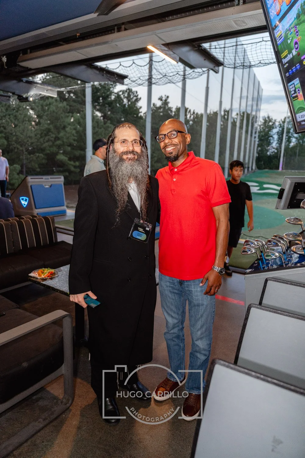 Two men standing together at a golf event, one wearing a black suit with a long beard and the other in a red polo shirt, with golf clubs and a screen showing a golf course in the background.