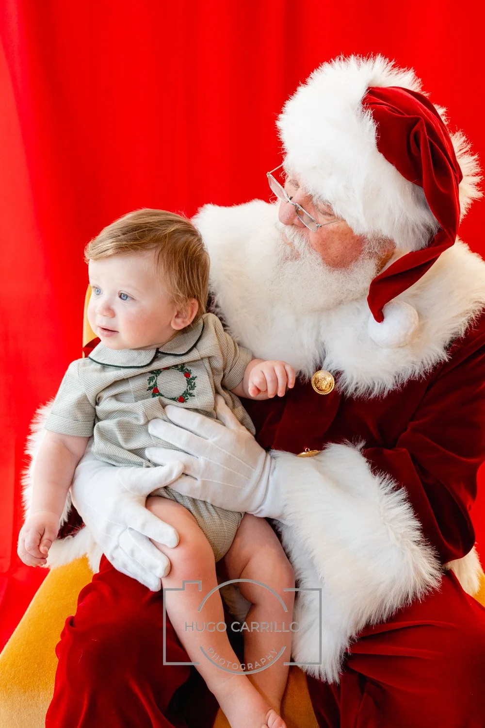 A young child sitting on Santa Claus's lap, looking to the side with a curious expression. Santa is wearing a red hat with white fur trim and a red velvet suit with gold buttons, with a red curtain backdrop.