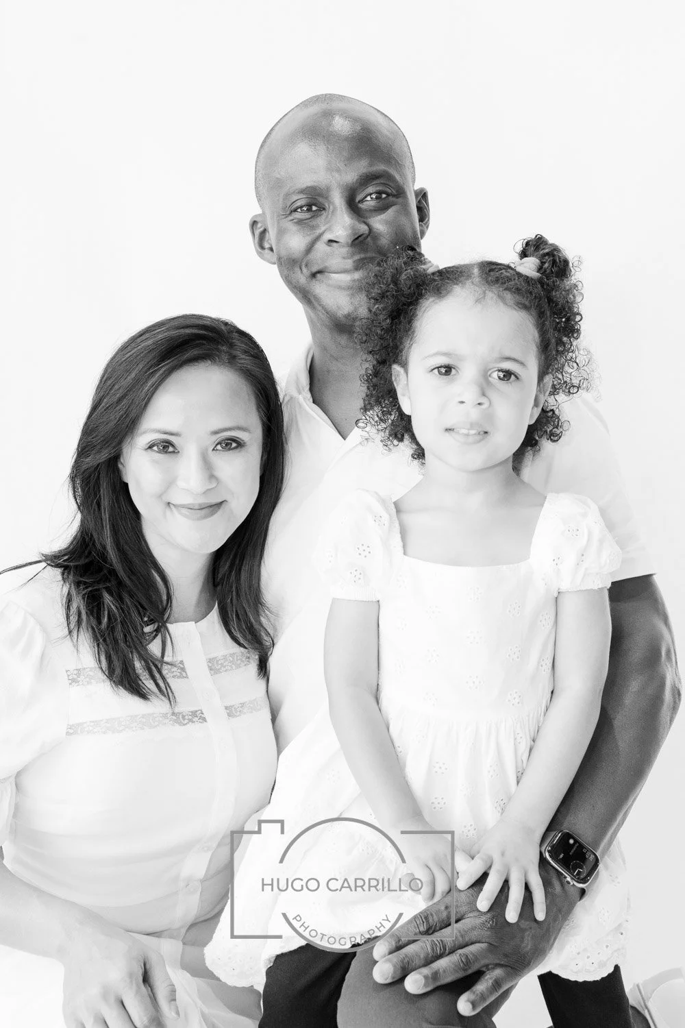 Black-and-white portrait of a family of three, including a woman, a man, and a young girl, against a plain white background. The woman has shoulder-length dark hair and is smiling, the man has a shaved head and a slight smile, and the young girl has 