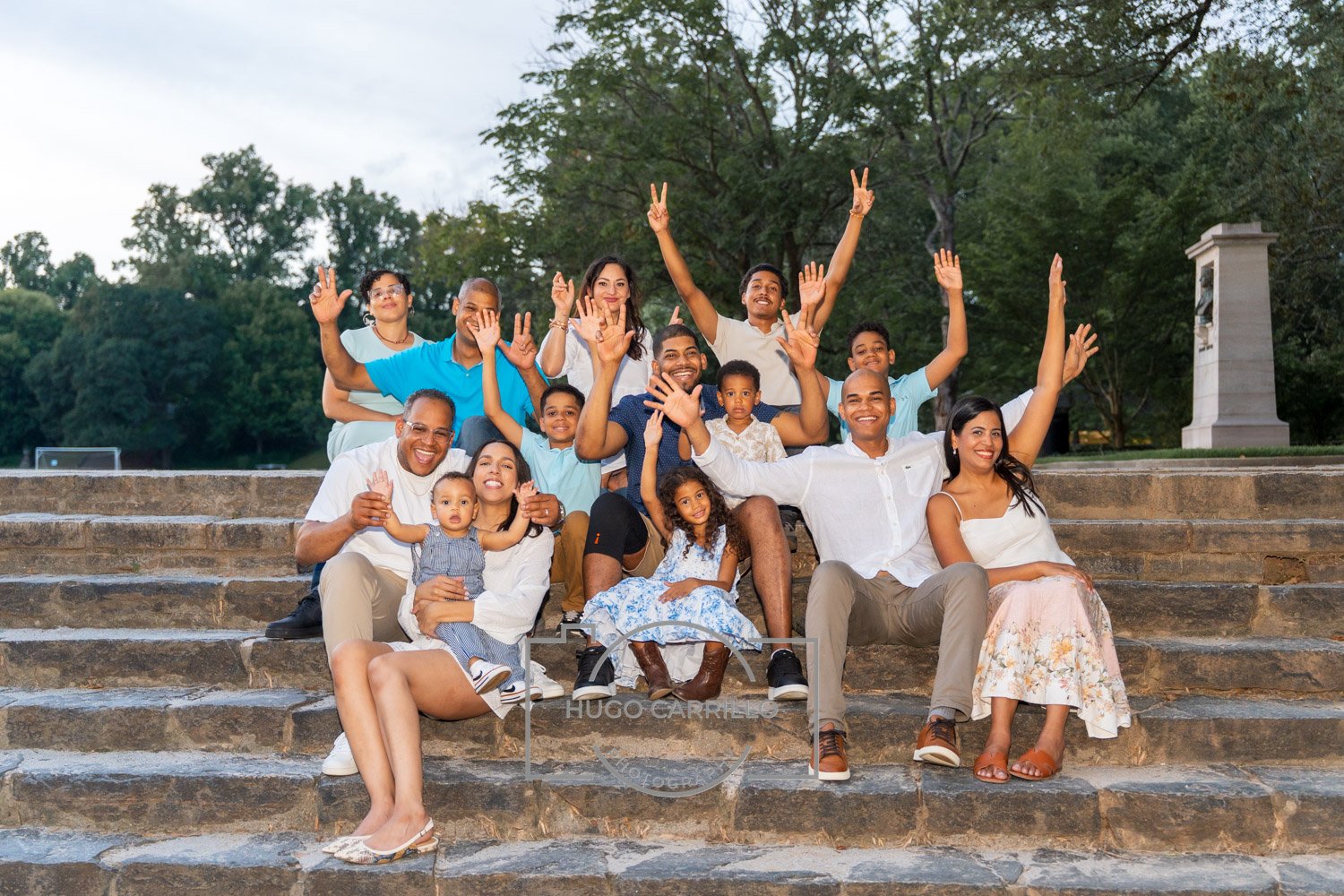 A diverse group of people, including children and adults, sitting and standing on stone steps outdoors, smiling, waving, and making peace signs in a park with trees in the background.