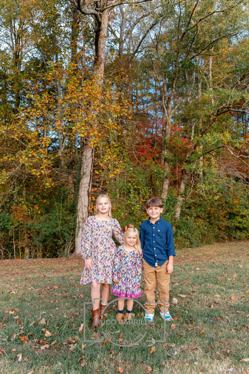 Three children standing outdoors in front of trees with autumn-colored leaves. The girl on the left wears a floral dress and brown boots, the girl in the middle wears a similar dress with pink accents, and the boy on the right wears a blue shirt and 