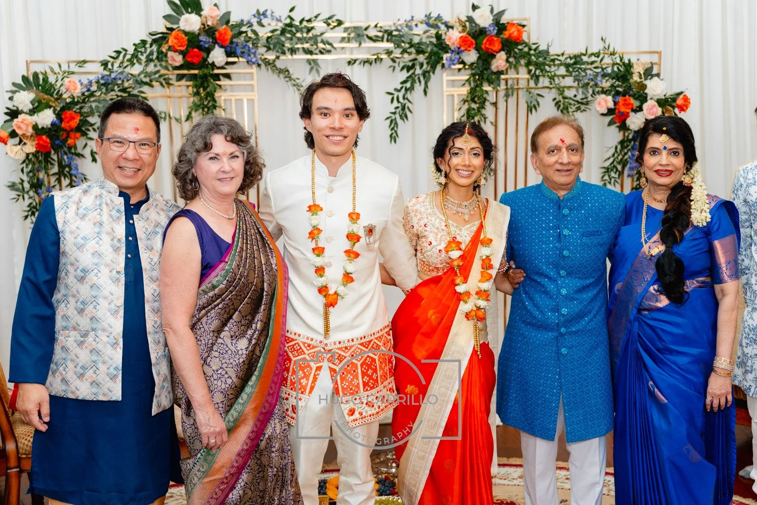 A group of seven people celebrating at a traditional Indian wedding, standing in front of floral decorations with white drapes. The bridal couple and their family members are dressed in colorful traditional Indian attire, wearing flower garlands and 