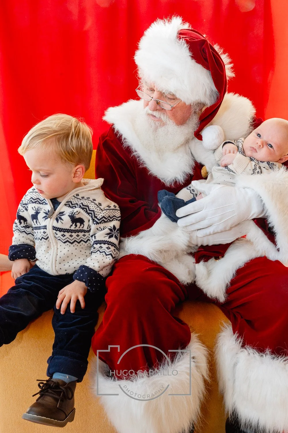 Santa Claus holding a baby and sitting next to a young boy in front of a red curtain.