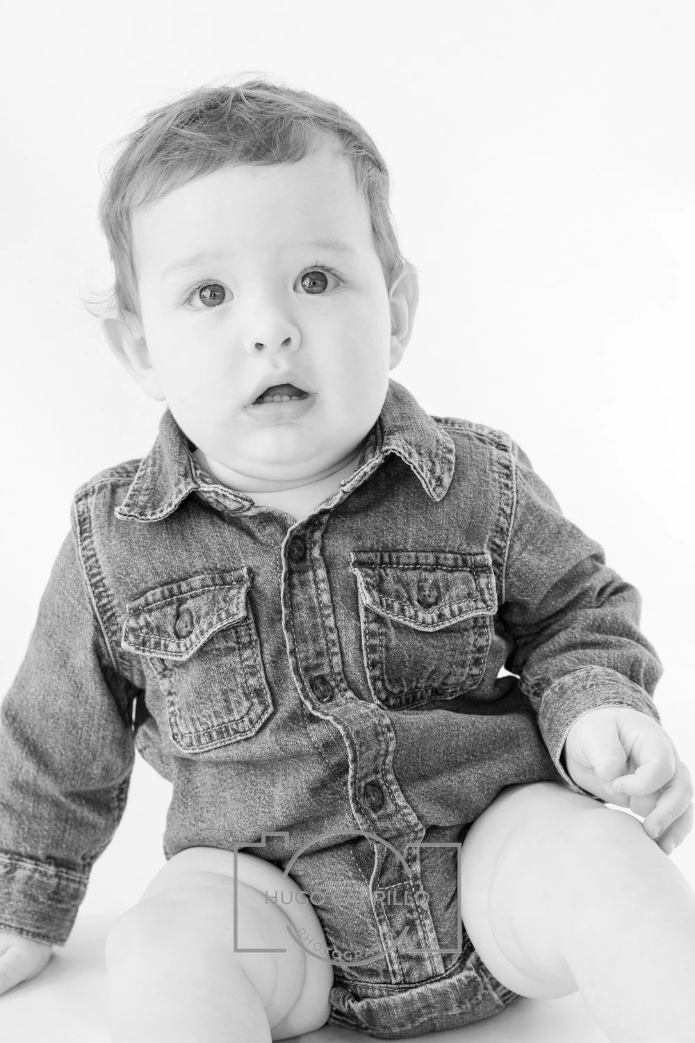 A black and white photograph of a young child with short hair, wearing a denim jacket, sitting on the floor with legs apart, looking slightly upward with a curious expression.