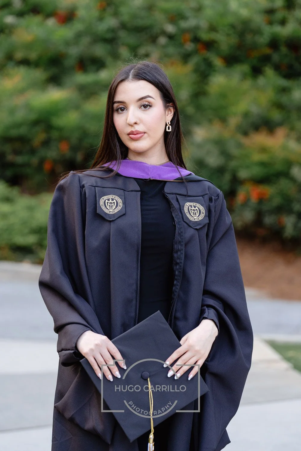 Young woman with long dark hair and earrings in a graduation gown and cap, holding the cap and standing outdoors with trees in the background.