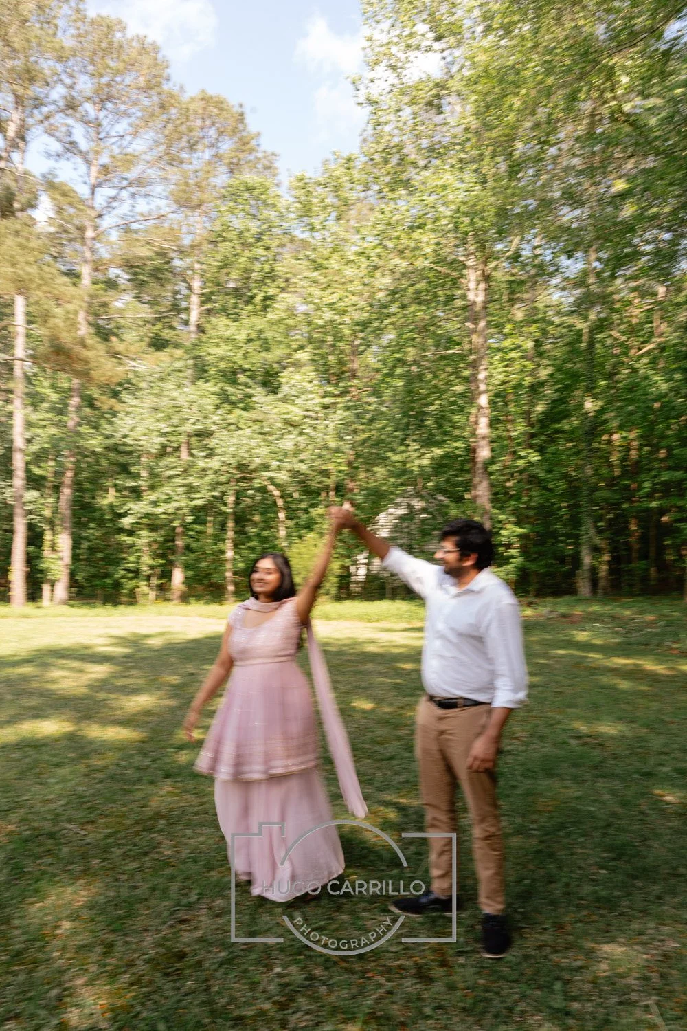 A couple dancing in a grassy, wooded park on a sunny day, with the woman in a pink dress and the man in a white shirt and tan pants.