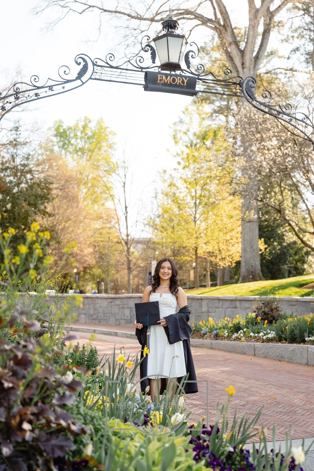 A young woman in a white dress holding a diploma, standing under an ornate sign that says "Emory," in a garden filled with flowers and trees, celebrating graduation.
