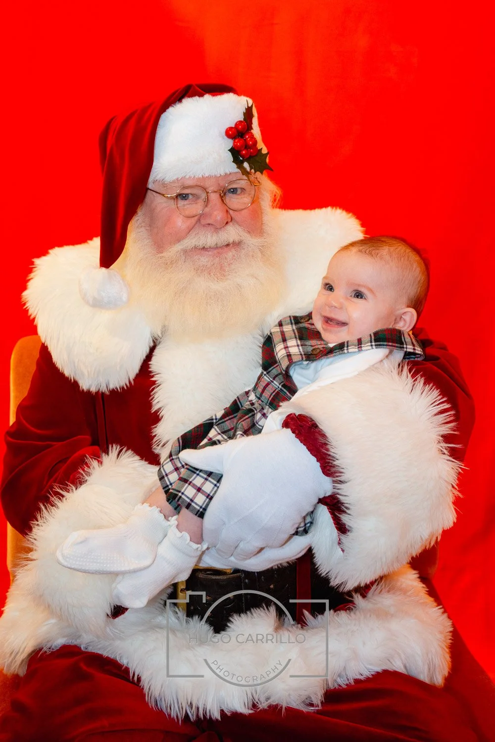 Santa Claus holding a smiling baby in front of a red background.