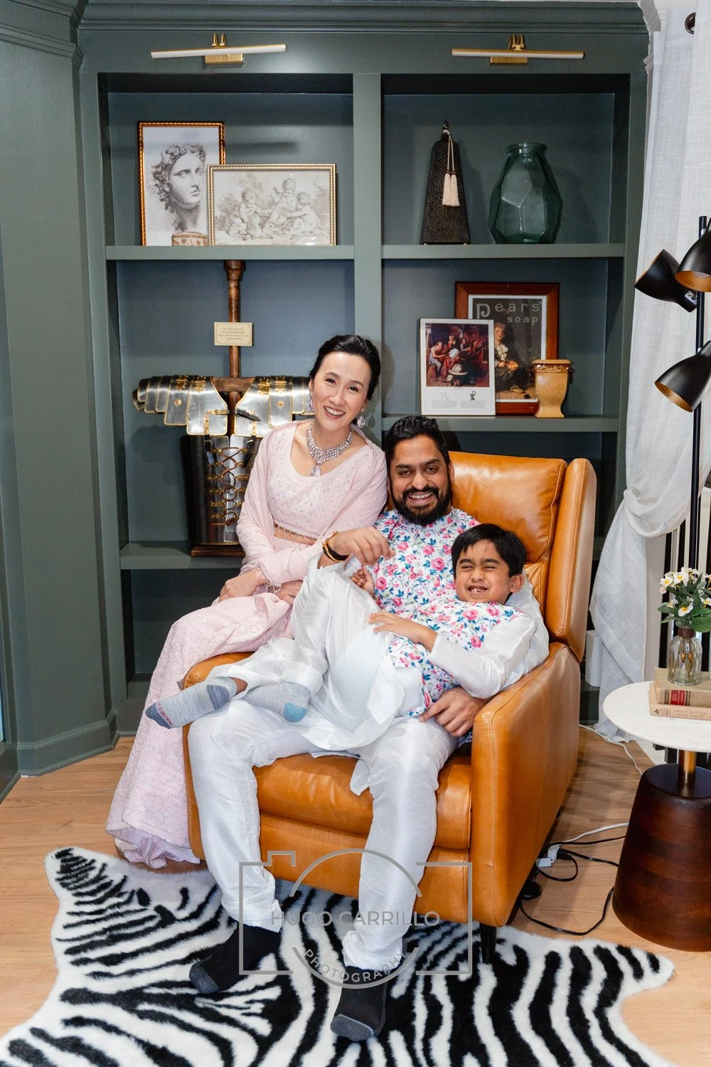 A family of three enjoying a moment together indoors, seated on a leather armchair with a black and white zebra-patterned rug beneath. The woman is dressed in a pink traditional outfit, the man is in white while the young boy is in a white shirt with
