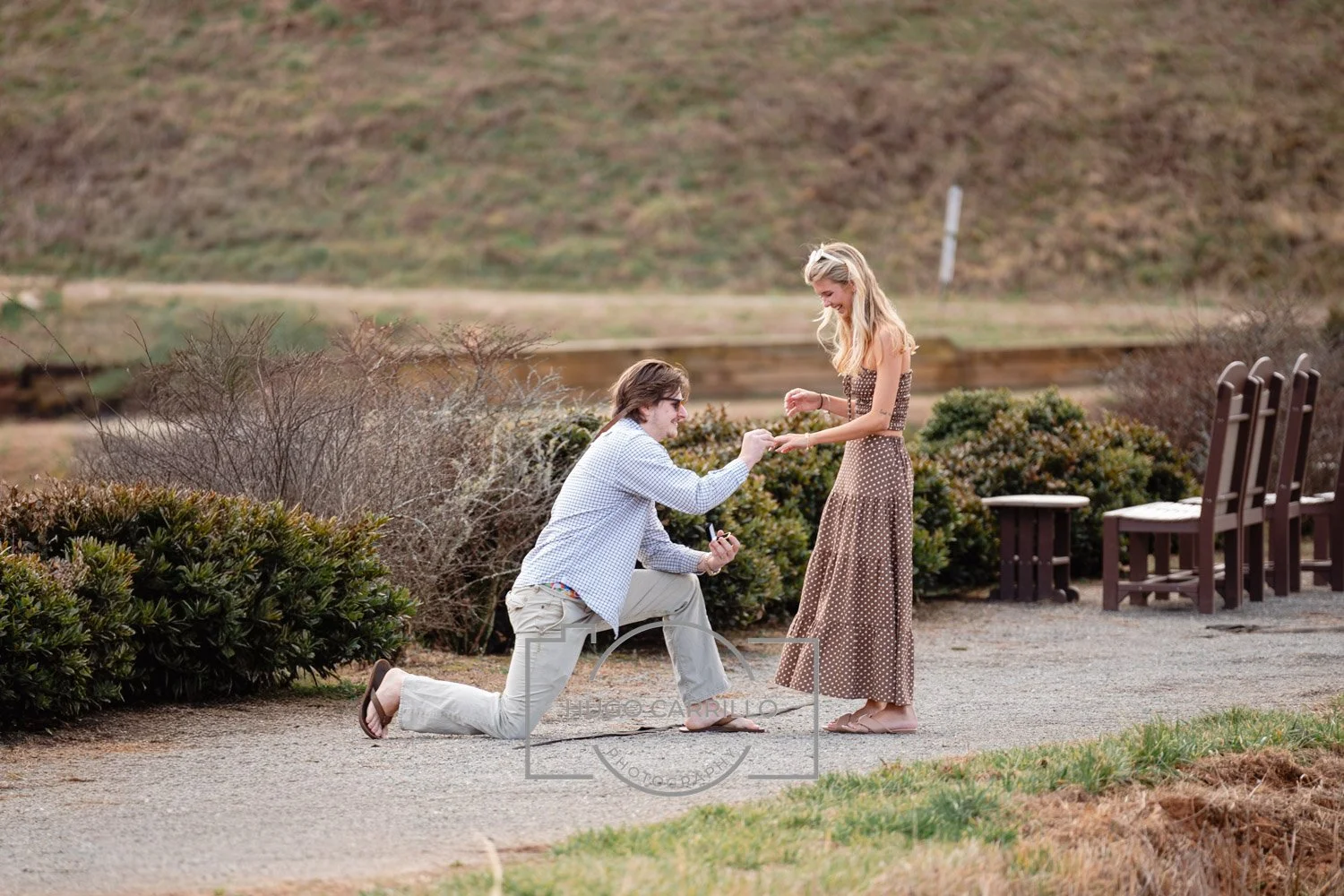 A man is proposing marriage to a woman outdoors in a park, with the man kneeling on one knee and holding a ring, while the woman looks pleasantly surprised.