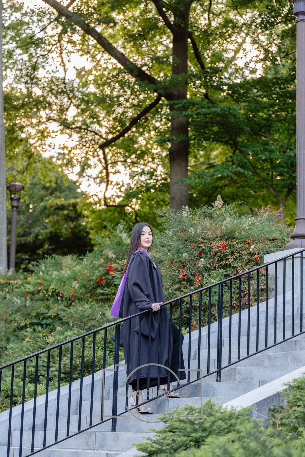 A woman in graduation regalia, wearing a black gown and a purple hood, stands on an outdoor staircase holding the railing, surrounded by greenery and trees, with warm sunlight filtering through the leaves.