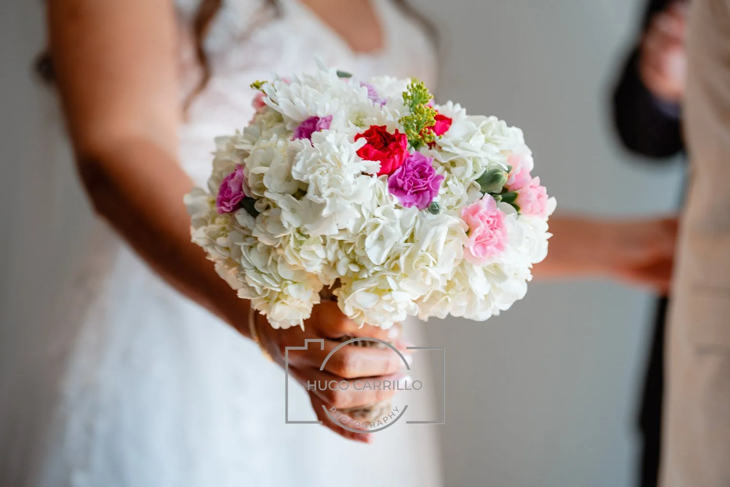 Woman in wedding dress holding a bouquet of white, pink, and purple flowers, with two blurred people in the background.