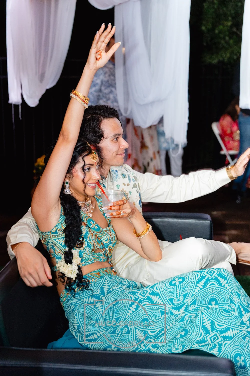 A couple, dressed in traditional Indian attire, sitting closely on a black chair at an outdoor event. The woman has long, dark curly hair with white flower accessories, and is wearing blue and gold jewelry and a patterned turquoise outfit. She is hol