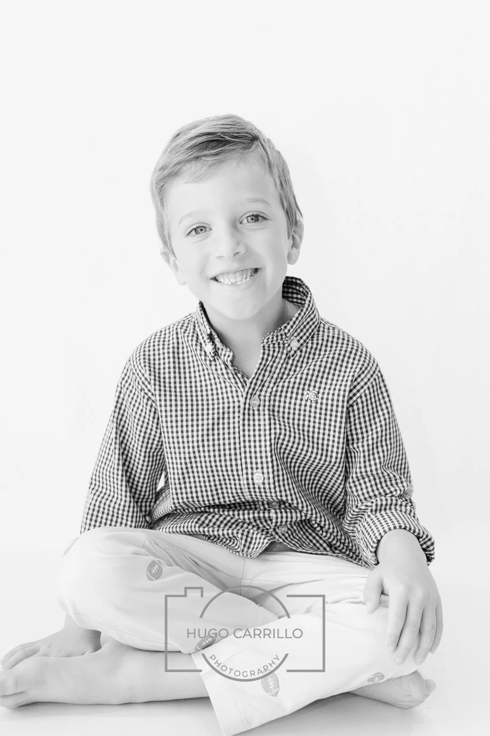 Black and white portrait of a smiling young boy sitting cross-legged on the floor, wearing a checkered shirt and patterned pants, against a plain background.