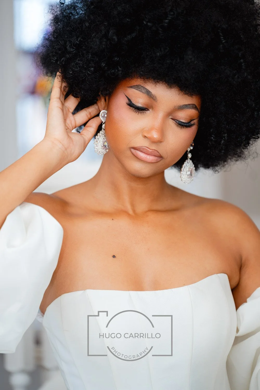 A woman with dark curly hair wearing a white off-shoulder dress and large earrings, adjusting her hair, with a neutral makeup look and closed eyes, in a well-lit indoor setting.