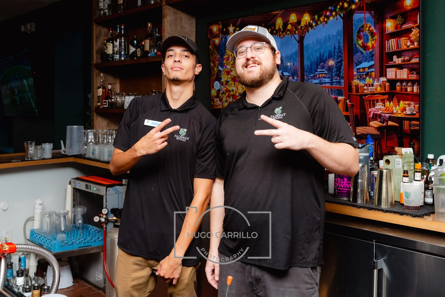 Two servers standing behind a bar, posing and making peace signs. They are wearing black shirts with a logo that says 'Fairway Social'. The bar has bottles, glasses, and bar equipment, with a festive Christmas holiday backdrop.