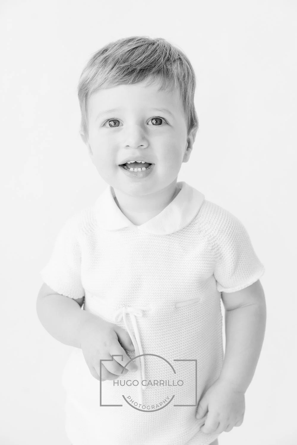 Black and white portrait of a smiling young boy with short hair, wearing a knitted top, standing against a plain background.