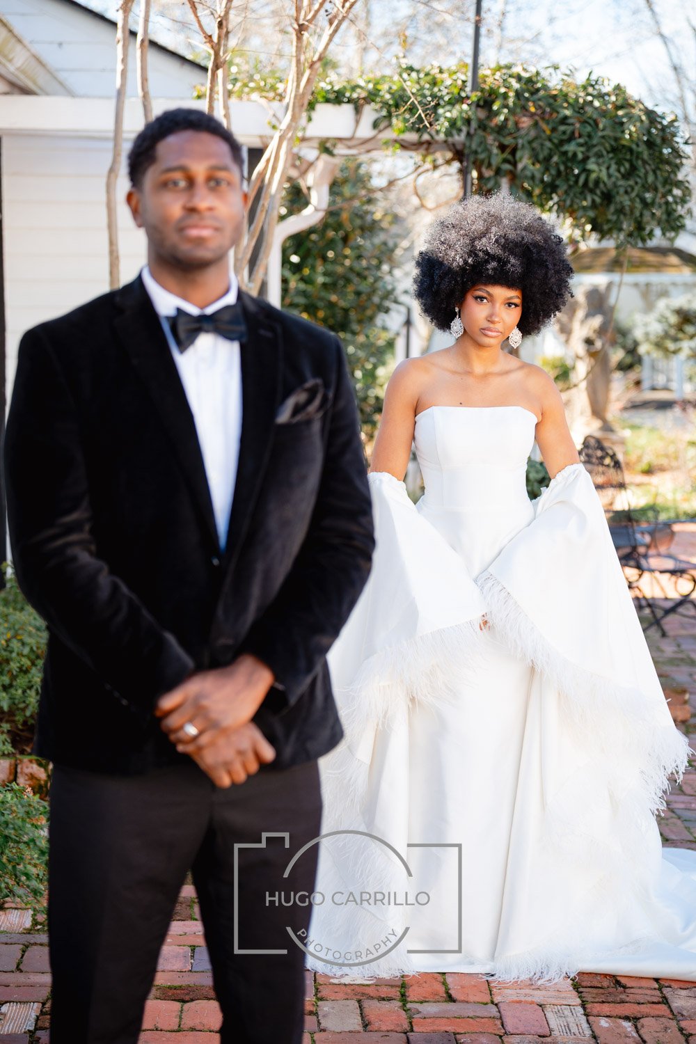 A woman in a white wedding gown with puffed long sleeves and large earrings, posing outdoors with a man in a black tuxedo and bowtie, in a garden setting with trees and greenery.