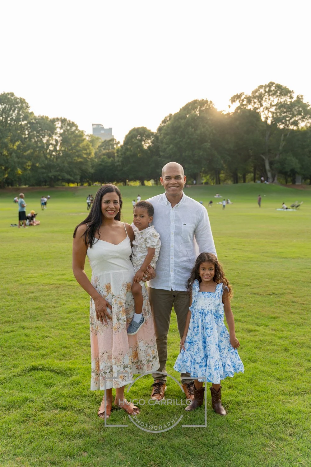 A family of four standing on a grassy field in a park, smiling at the camera during sunset. The mother is holding a young child, the father stands beside a young girl, and there are people in the background enjoying the park.
